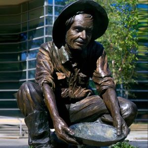 Bronze statue of an old man wearing a beret, rolled up sleeves, and short boots, squatting on the street pouring water with a basin