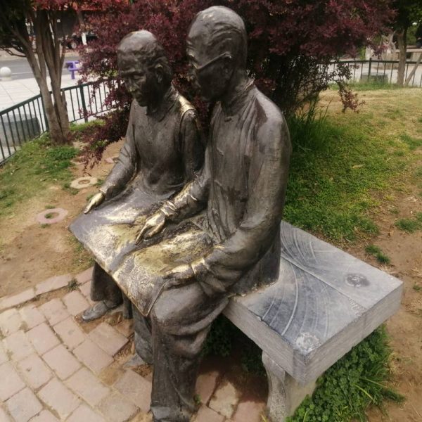 Bronze sculpture in an outdoor park showing two elderly people sitting side by side on a stone bench - Image 3