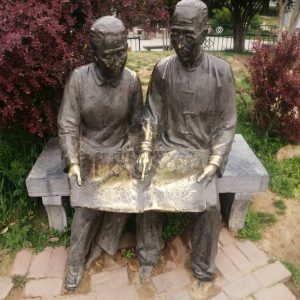 Bronze sculpture in an outdoor park showing two elderly people sitting side by side on a stone bench